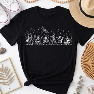 May include: A black short-sleeved t-shirt featuring a white line art graphic of various insects playing musical instruments, including a dragonfly, a ladybug, and a bee, accompanied by musical notes, stars, and a crescent moon design.