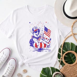 Könnte beinhalten: Ein weißes T-Shirt mit einer Grafik eines Hundes, der eine Sonnenbrille und eine Kappe mit der Aufschrift USA 280 trägt, auf einem rot-weiß-blau gestreiften Schwimmring sitzt, eine Flasche hält und eine US-Flagge schwenkt, umgeben von Feuerwerk.
