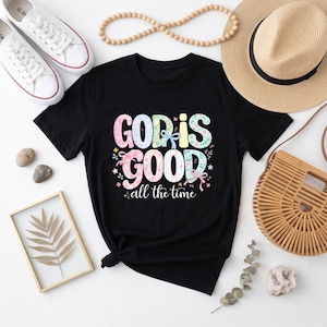 May include: A black short-sleeved t-shirt featuring the colourful text "GOD IS GOOD all the time" in a decorative font with floral and ribbon accents. The shirt is surrounded by white trainers, a straw hat, a wooden bag, and a framed leaf print.