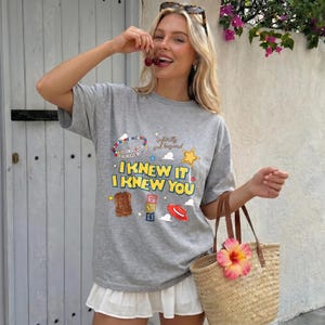 K&ouml;nnte beinhalten: Ein graues, kurz&auml;rmeliges T-Shirt mit einem Grafikdruck, der den Text I KNEW IT I KNEW YOU, infinity and beyond sowie Illustrationen eines Perlenarmbands, eines braunen Cowboystiefels, von Buchstabenw&uuml;rfeln, eines roten Hutes und eines gelben Sterns zeigt.