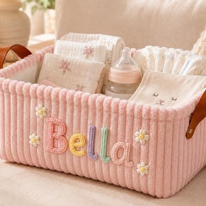 May include: A rectangular pink corduroy storage basket featuring the name Bella embroidered in pastel letters on the front, accented with small white daisy appliques and brown leather side handles, containing baby cloths and a plastic baby bottle.