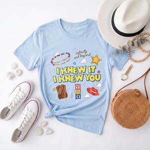 K&ouml;nnte beinhalten: Ein hellblaues Kurzarm-T-Shirt mit dem Text "I KNEW IT I KNEW YOU" in Gelb mit blauen Umrandungen. Die Grafik zeigt ein Perlenarmband, einen Sheriffstern, Cowboystiefel, Spielzeugw&uuml;rfel, einen roten Hut und den Schriftzug "infinity and beyond".