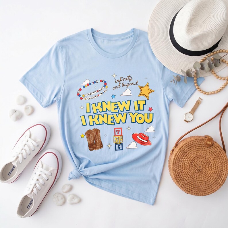 K&ouml;nnte beinhalten: Ein hellblaues Kurzarm-T-Shirt mit dem Text "I KNEW IT I KNEW YOU" in Gelb mit blauen Umrandungen. Die Grafik zeigt ein Perlenarmband, einen Sheriffstern, Cowboystiefel, Spielzeugw&uuml;rfel, einen roten Hut und den Schriftzug "infinity and beyond".