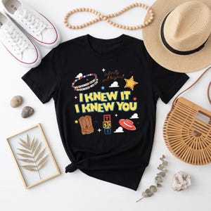 K&ouml;nnte beinhalten: Ein schwarzes Kurzarm-T-Shirt mit einem Grafikdruck mit dem Text "I KNEW IT I KNEW YOU" und "infinity and beyond". Das Design enth&auml;lt Illustrationen eines Perlenarmbands, eines gelben Sterns, brauner Cowboystiefel, Buchstabenw&uuml;rfel und eines roten Hutes.
