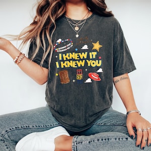 K&ouml;nnte beinhalten: Ein anthrazitfarbenes Kurzarm-T-Shirt mit dem gelben Schriftzug I KNEW IT I KNEW YOU. Die Grafik zeigt ein buntes Perlenarmband, einen gelben Sheriffstern, einen braunen Cowboystiefel, gestapelte Buchstabenw&uuml;rfel und einen roten Cowboyhut.