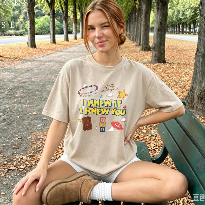 K&ouml;nnte beinhalten: Ein beiges kurz&auml;rmeliges T-Shirt mit einem Grafikdruck mit dem Text I KNEW IT I KNEW YOU, infinity and beyond, einem Perlenarmband, einem gelben Stern, einem braunen Cowboystiefel, gestapelten Alphabetw&uuml;rfeln und einem roten Cowboyhut.
