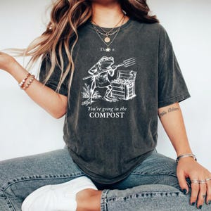 Könnte beinhalten: Ein anthrazitfarbenes Kurzarm-T-Shirt mit einer weißen Grafik eines Frosches, der eine Heugabel neben einer Holzkiste hält. Der Text "That's it." steht über dem Frosch und "You're going in the COMPOST" unter der Grafik in weißer Schrift.