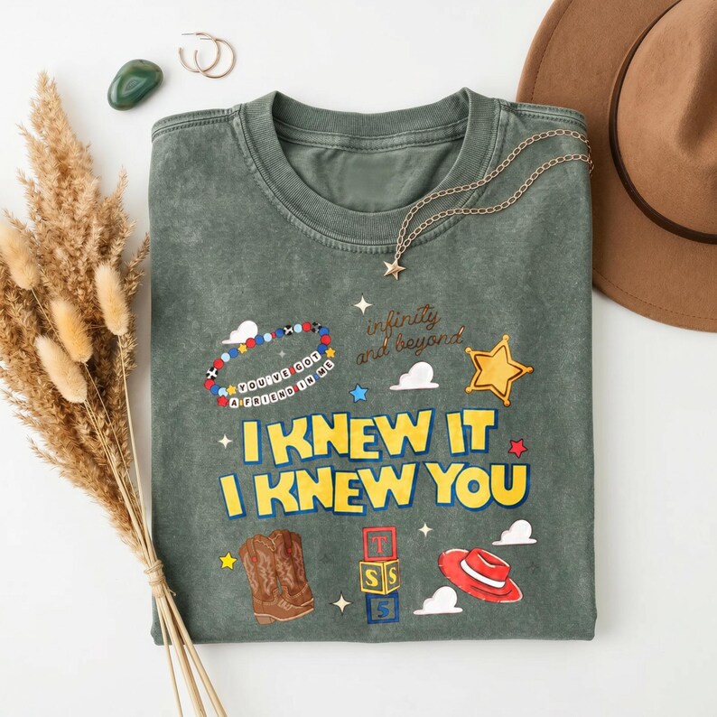 K&ouml;nnte beinhalten: Ein dunkelolivgr&uuml;nes, mineralgewaschenes T-Shirt mit einem Grafikdruck, der den Text "I KNEW IT I KNEW YOU", "infinity and beyond" und "YOU'VE GOT A FRIEND IN ME" enth&auml;lt. Das Design zeigt ein Perlenarmband, einen gelben Stern, einen braunen Cowboystiefel, Buchstabenw&uuml;rfel und einen roten Hut.