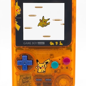 Może przedstawiać: Pomarańczowa konsola Game Boy Pocket z motywem Pikachu. Ekran wyświetla spikselowaną postać Pikachu w grze side-scrolling. Posiada niebieski d-pad, różowe i turkusowe przyciski oraz logo Pikachu.