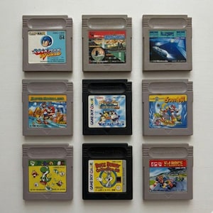 Puede incluir: Nueve cartuchos de juegos de Game Boy vintage dispuestos en una cuadrícula. Los cartuchos presentan etiquetas coloridas con títulos de juegos como "Rockman World", "Super Mario Land" y "Bugs Bunny Collection". Los cartuchos son grises y negros.