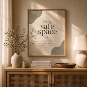May include: A framed print with the words "THIS IS A safe space" in a soft, neutral colour palette. The print includes the phrases "YOU ARE SEEN, YOU ARE HEARD, YOU ARE VALUED, YOU ARE WELCOME HERE." The print is displayed above a wooden cabinet.