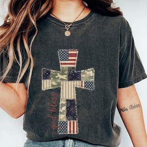 May include: Dark grey t-shirt with a patchwork cross design. The cross incorporates elements of the American flag, camouflage, and striped patterns. The words "god bless" are written vertically on the left side of the cross in a cursive font.