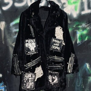 Puede incluir: Un abrigo de piel sintética negra con varios parches, incluyendo uno que dice PUNX NUEVOS GUERRERO y otro que dice PUNKS NOT DEAD. La prenda está decorada con múltiples cremalleras metálicas plateadas, imperdibles y trozos de tela rasgada.
