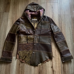 Puede incluir: Una chaqueta de cuero con capucha que presenta un diseño de parches en varios tonos marrones y oliva. La parte frontal muestra una textura con patrón de llamas en el lado izquierdo, cierre de botones con herrajes metálicos y forro morado.
