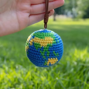 Può includere: Ornamento a forma di globo terrestre all'uncinetto con oceani blu, masse terrestri verdi e continenti gialli. Sospeso da un cordino marrone. Sfondo verde sfocato.