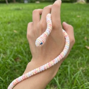 Puede incluir: Un juguete de serpiente de ganchillo en colores pastel rosa, amarillo y blanco. La cabeza de la serpiente tiene un ojo azul. La serpiente está envuelta alrededor de una mano, con un fondo de hierba verde.