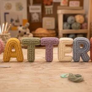 Op de afbeelding: Een set van zeven driedimensionale gehaakte letters die het woord PATTERN vormen. Elke letter heeft een eigen kleur: roze, geel, saliegroen, lavendel, crème, lichtblauw en koraal, allemaal vervaardigd met een zichtbare stekenstructuur.