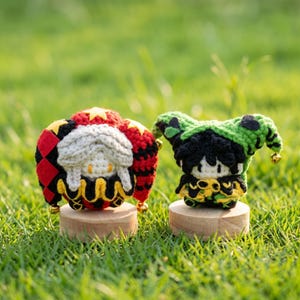 May include: Two handmade crochet jester dolls. One doll features a red, black, and yellow color scheme, while the other is green, black, and yellow. Both dolls are mounted on small wooden bases and have small gold bells.