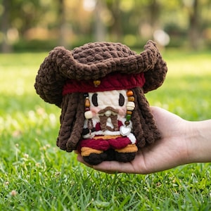May include: A handmade crochet doll resembling a pirate, featuring a brown hat, red bandana, and detailed facial features. The doll has brown dreadlocks, a white shirt, and a red and gold vest. It is held in a person's hand against a blurred green background.