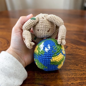 Op de afbeelding: Een handgemaakt gehaakt speelgoed met een beige figuur die een wereldbol omarmt. De wereldbol is een bal met blauwe oceanen, groene en gele continenten. Een uniek, milieuvriendelijk item.