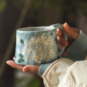 May include: A light blue ceramic mug with a textured, uneven surface featuring hand-painted white hydrangea clusters with small yellow centers and green leaves. The mug has a curved handle and a slightly flared rim, showcasing a rustic finish.