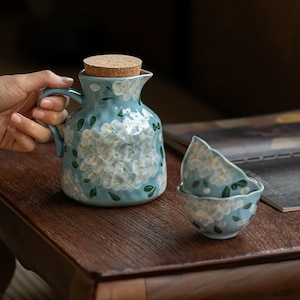 May include: A light blue ceramic pitcher with a cork stopper and two matching small bowls, all decorated with hand-painted white hydrangea flowers and green leaves. The pitcher features a curved handle and a textured, uneven surface finish.