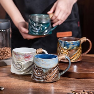 May include: A collection of handmade ceramic mugs with unique designs and rope-wrapped handles. The mugs feature a variety of colors, including blue, brown, and white, and are displayed on a wooden surface. The mugs are approximately 4 inches tall.
