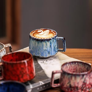 May include: A blue and white ceramic mug with a handle, filled with coffee and latte art. The mug has a textured, glazed finish. Other mugs in red and blue are visible in the foreground.