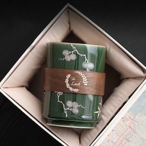 May include: A cylindrical green ceramic cup featuring white botanical illustrations of pine needles and branches. A dark brown wooden band wraps around the center, displaying the text The Luck enclosed within a white circular leaf wreath design.