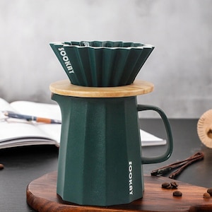 May include: A dark green coffee pour-over set with a wooden lid and a matching carafe. The filter holder has a pleated design and the word "SOOKAY" printed on it. The carafe has a handle and a geometric shape. Coffee beans are scattered on the wooden surface.