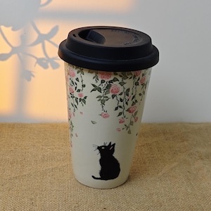 May include: A white travel mug with a black lid and a black cat illustration. The mug is decorated with pink roses and green leaves. The mug is approximately 6 inches tall.