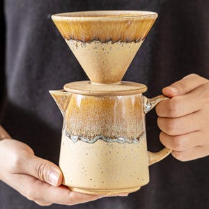 Peut inclure: Un ensemble de cafeti&egrave;re &agrave; verser en c&eacute;ramique avec une palette de couleurs chaudes et terreuses. L'ensemble comprend un verseur en forme de c&ocirc;ne, un couvercle et une petite carafe avec une poign&eacute;e. Le design pr&eacute;sente une finition glac&eacute;e tachet&eacute;e avec des tons bruns et cr&egrave;me.
