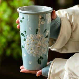 May include: A tall, cylindrical ceramic tumbler in a light blue glaze, featuring a hand-painted design of white hydrangea blossoms with small yellow centers and dark green leaves. The surface has a glossy finish and a smooth, tapered shape.
