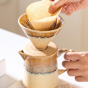 Peut inclure: Un ensemble de cafeti&egrave;re &agrave; verser en c&eacute;ramique avec un gla&ccedil;age marron et beige. Une main ins&egrave;re un filtre en papier dans le verseur en forme de c&ocirc;ne. La cafeti&egrave;re a un couvercle et une poign&eacute;e. Le design est rustique et artisanal.