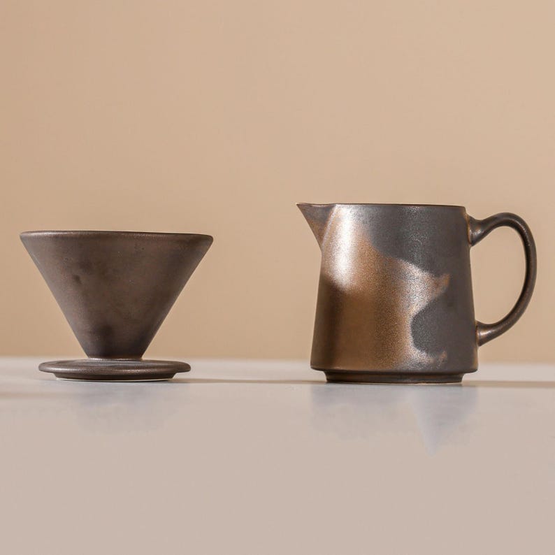 Peut inclure: Un ensemble de cafeti&egrave;re &agrave; verser et de pichet en c&eacute;ramique brun fonc&eacute;. La cafeti&egrave;re a une forme conique et repose sur une petite base. Le pichet a une anse et un bec verseur. Les deux pi&egrave;ces ont un &eacute;clat m&eacute;tallique.