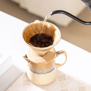 Peut inclure: Une cafeti&egrave;re &agrave; verser en c&eacute;ramique avec un gla&ccedil;age beige et marron. De l'eau chaude est vers&eacute;e sur du caf&eacute; moulu dans un filtre en papier. La cafeti&egrave;re a une poign&eacute;e et un bec verseur.