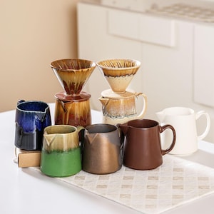 Peut inclure: Ensemble de cafeti&egrave;res &agrave; verser et de tasses en c&eacute;ramique. Les cafeti&egrave;res comprennent un porte-filtre conique et une carafe assortie. Les tasses sont de diff&eacute;rentes couleurs, dont le bleu, le vert, le marron et le blanc. Les articles sont dispos&eacute;s sur une surface blanche.