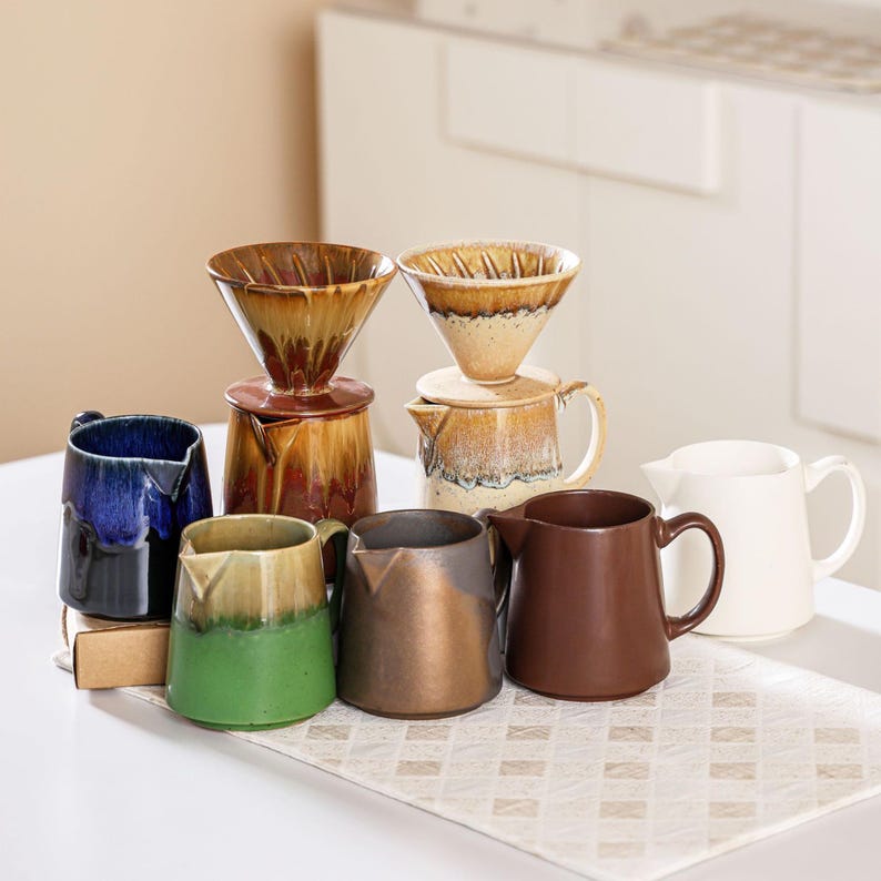 Peut inclure: Ensemble de cafeti&egrave;res &agrave; verser et de tasses en c&eacute;ramique. Les cafeti&egrave;res comprennent un porte-filtre conique et une carafe assortie. Les tasses sont de diff&eacute;rentes couleurs, dont le bleu, le vert, le marron et le blanc. Les articles sont dispos&eacute;s sur une surface blanche.