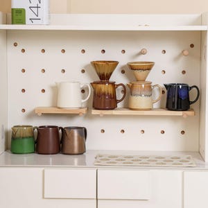 Peut inclure: Un meuble blanc avec un fond en panneau perfor&eacute; pr&eacute;sente diverses cafeti&egrave;res en c&eacute;ramique, des c&ocirc;nes &agrave; verser et de petits pichets. Les articles sont dans des tons de marron, beige, vert et bleu. Des &eacute;tag&egrave;res en bois et un tapis &agrave; motifs compl&egrave;tent l'ensemble.