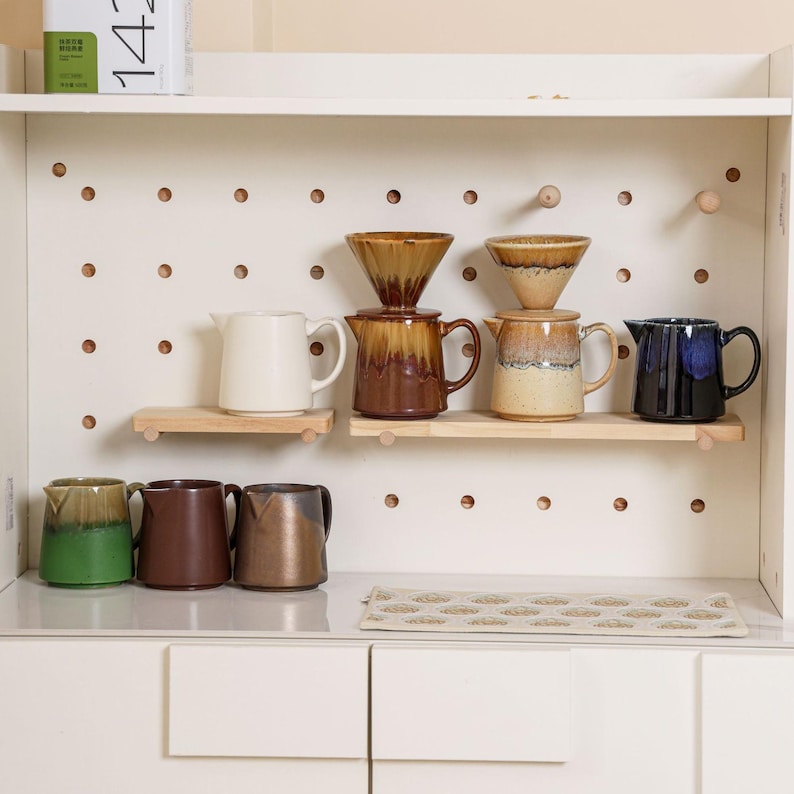 Peut inclure: Un meuble blanc avec un fond en panneau perfor&eacute; pr&eacute;sente diverses cafeti&egrave;res en c&eacute;ramique, des c&ocirc;nes &agrave; verser et de petits pichets. Les articles sont dans des tons de marron, beige, vert et bleu. Des &eacute;tag&egrave;res en bois et un tapis &agrave; motifs compl&egrave;tent l'ensemble.