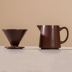 Peut inclure: Un ensemble de cafeti&egrave;re &agrave; verser et de pichet en c&eacute;ramique brun fonc&eacute;. Le syst&egrave;me de versement a une forme conique et repose sur une base circulaire. Le pichet a une poign&eacute;e et un bec verseur.