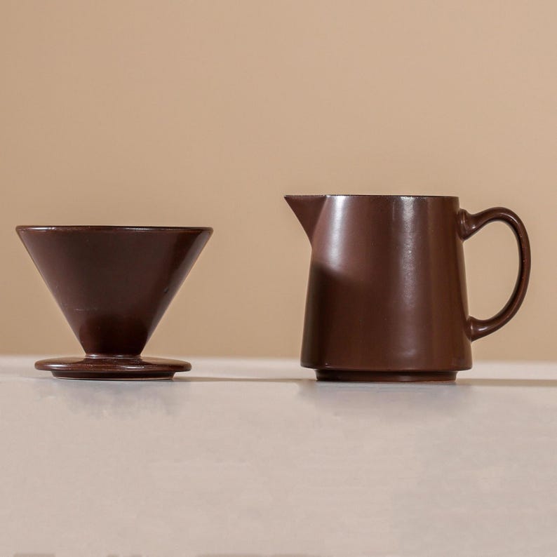 Peut inclure: Un ensemble de cafeti&egrave;re &agrave; verser et de pichet en c&eacute;ramique brun fonc&eacute;. Le syst&egrave;me de versement a une forme conique et repose sur une base circulaire. Le pichet a une poign&eacute;e et un bec verseur.