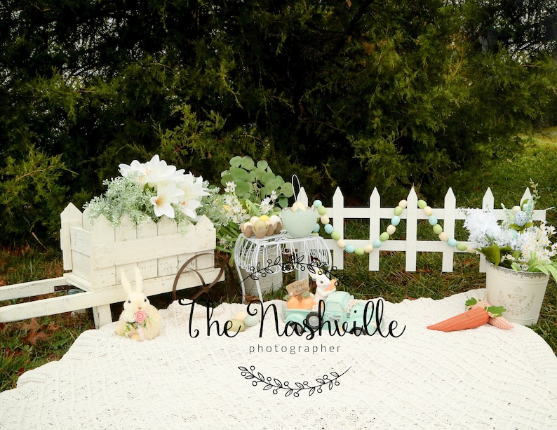 May include: A white wooden wagon with a white picket fence and pastel colored pom poms. The wagon is filled with white flowers and there is a white bunny rabbit sitting in front of it. There is a white crocheted blanket on the ground with a carrot and a blue toy car. The text "The Nashville photographer" is written on the blanket.