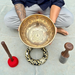 May include: A gold-toned metal singing bowl featuring intricate etched Sanskrit mantras and Om symbols, resting on a black and gold patterned fabric cushion, accompanied by a wooden mallet with a red felt tip and a separate wooden striker.