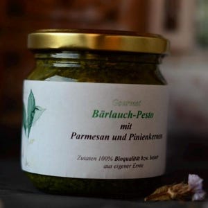 Könnte beinhalten: Ein Glas Gourmet-Bärlauch-Pesto mit Parmesan und Pinienkernen. Das Etikett enthält deutschen Text. Das Glas hat einen goldenen Deckel und ein weißes Etikett mit grünem Text und einer Blattillustration. Das Pesto ist leuchtend grün.