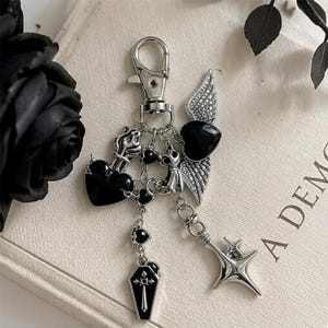 May include: A silver-toned metal keychain featuring multiple charms including a black heart, a black heart with silver wings, a small black coffin with a cross design, and a four-pointed star. The charms are attached to a silver lobster clasp.