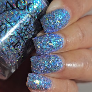 May include: Close-up of manicured nails with a sparkling, iridescent blue glitter polish. The polish reflects multiple colors, creating a shimmering effect. The bottle of nail polish is partially visible, with the text "MAGIC" and a spiderweb design.
