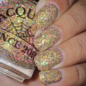 Puede incluir: Primer plano de una mano con esmalte de uñas con purpurina. Las uñas están cubiertas de purpurina brillante e iridiscente que refleja tonos dorados, verdes y rosas. Una botella de esmalte de uñas es parcialmente visible, con el texto "AQUANCE 2011" y un diseño de telaraña.