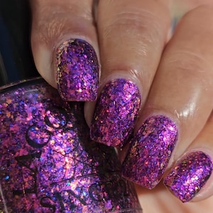 May include: Close-up of a hand with manicured nails. The nails are painted with a vibrant purple glitter polish, reflecting light. The polish has a textured appearance with gold and iridescent flakes.