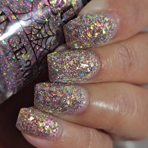May include: Close-up of a hand with manicured nails covered in iridescent glitter. The glitter is a mix of pink, purple, gold, and green. A bottle of nail polish is partially visible in the background, with a spiderweb design.